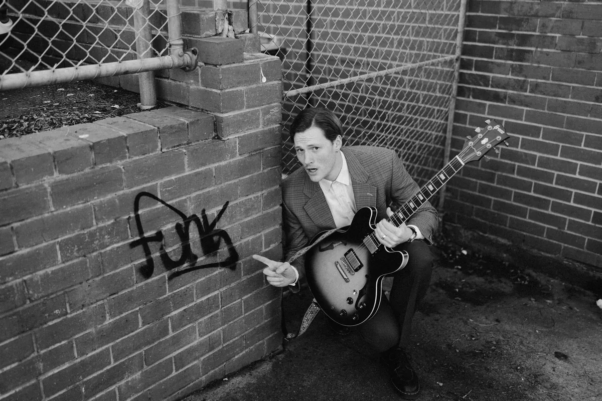 Jem Pryse crouched against a brick wall with a guitar